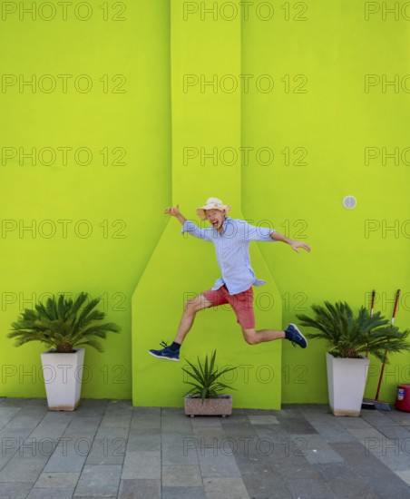 Young man jumping over plant pot in front of green house wall, green house facade with flowers, colourful houses on the island of Burano, Venice, Veneto, Italy