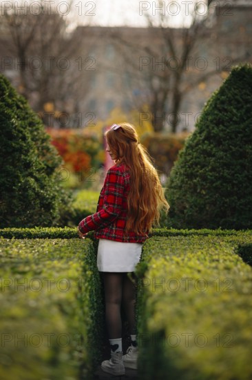 A young woman with long red hair explores a lush green maze in a Montreal garden during fall. Vibrant autumn colors and urban background create a serene atmosphere