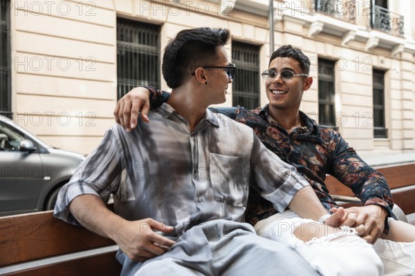 A cheerful latin gay couple sits close together on a bench, laughing and enjoying a sunny day in the vibrant streets of Madrid, showcasing love and happiness