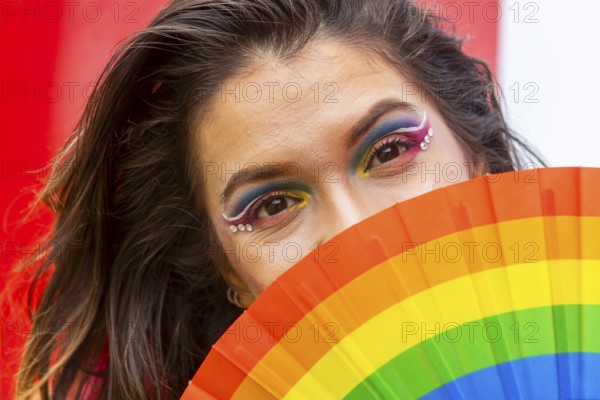A woman with vibrant rainbow makeup partially conceals her face with a pride-themed fan. Her expressive eyes convey the spirit of inclusion, celebrating LGBTQIA+ identity