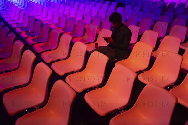 A indian man sits among vibrant, colorful auditorium chairs at a conference of AI in Amsterdam engrossed in his smartphone. The scene contrasts solitary focus against a backdrop of empty seats