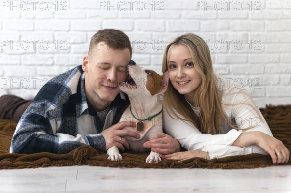 A young couple shares a loving moment with their energetic white and brown dog, lying on a cozy blanket at home, smiling