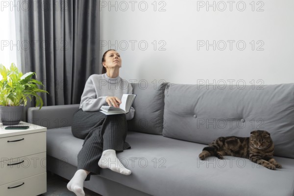 A woman sits on a gray sofa, holding a book, looking peaceful and contemplative. Beside her, a cat lounges calmly. A green plant and drawer add to the room's tranquility
