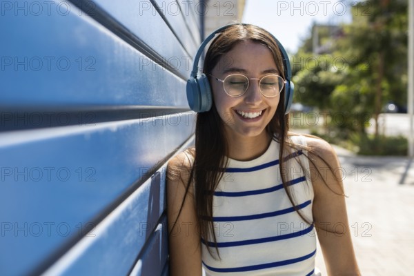 Young woman enjoying her urban lifestyle outdoors, leaning against a blue wall while listening to music through headphones. Her eyes are closed, and she is smiling peacefully