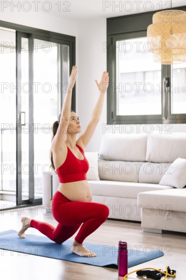 A pregnant woman practices yoga on a mat in a cozy living room, focusing on wellness and relaxation. She wears a red outfit and is surrounded by bright natural light