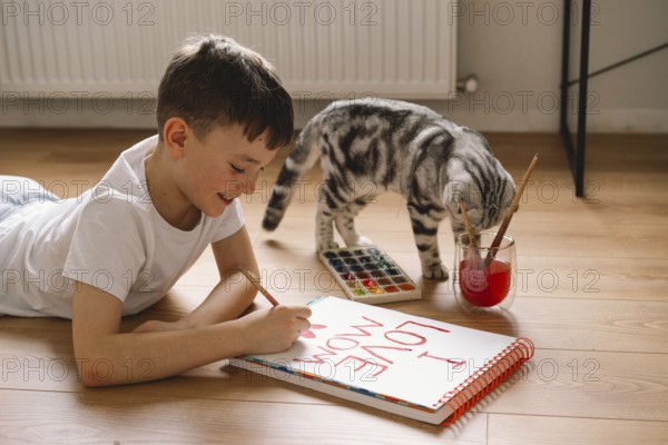 A boy lies on the floor, drawing a Mother's Day card with vibrant paints. His dedicated expression and playful cat create a heartwarming scene of creativity and love