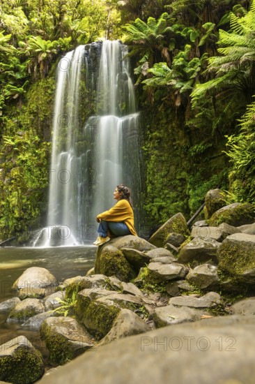 A woman sits on rocks, meditating beside a serene waterfall, surrounded by lush greenery in the Great Ocean Road, Australia. The tranquil scene embodies peace, reflection, and connection with nature