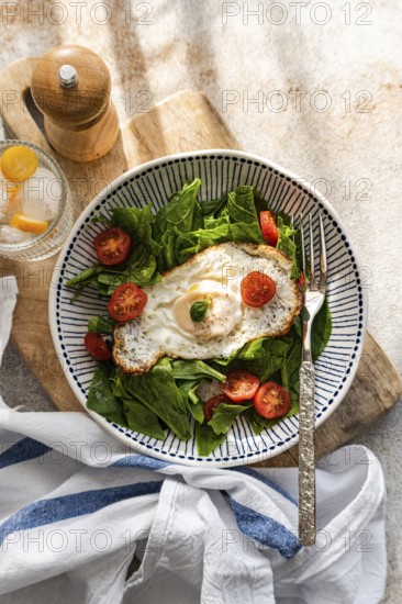 Top view of a fresh and nutritious lunch featuring crisp spinach leaves, juicy cherry tomatoes, and a perfectly fried egg, complemented by a refreshing glass of mineral water with ice and slices of kumquat