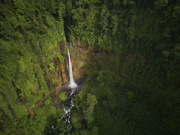 Aerial shot capturing the majestic Kaspa Biru Waterfall descending into a natural gorge, surrounded by dense tropical rainforest. The waterfall creates a serene atmosphere as it flows through the verdant landscape
