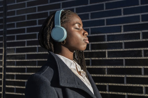 An African American businesswoman with braids enjoys a break, listening to music through headphones, eyes closed in relaxation, leaning against a brick wall outdoors