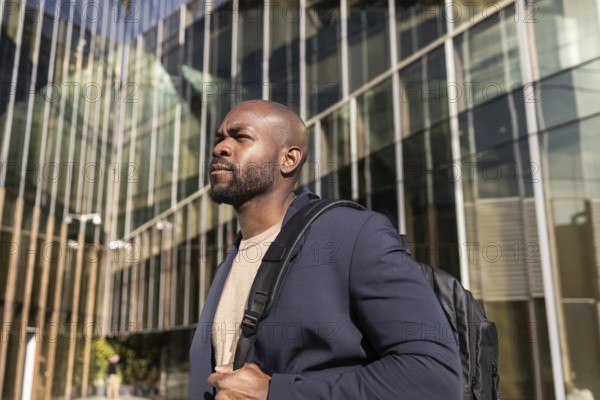 A confident cuban man with a backpack stands outside a modern office building. The glass facade reflects urban surroundings, creating a dynamic city atmosphere
