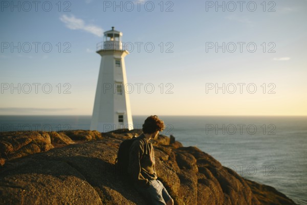 Side view of unrecognizable young man sitting on rocks gazing at the ocean near Cape Spear Lighthouse at sunset. The serene environment highlights a tranquil moment at this historic site in Newfoundland & Labrador, Canada