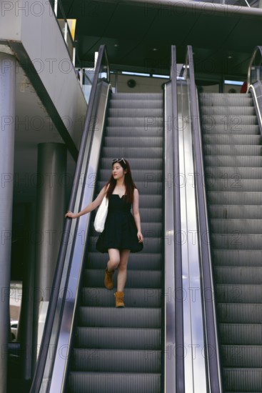A young asian woman in a black dress and boots descends an escalator in an urban setting. She carries a white bag and sunglasses, exuding casual style and confidence