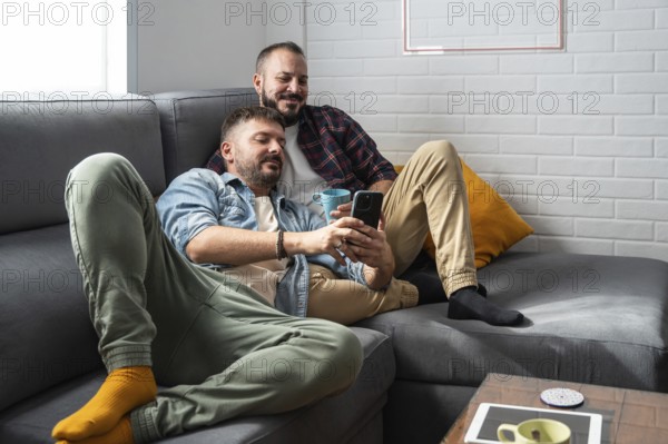 A content gay couple lounges on a comfortable gray sofa, enjoying coffee together while browsing a smartphone. They rest casually against soft pillows, sharing a peaceful moment