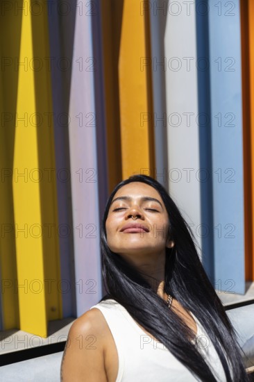 A woman in a colorful outfit enjoys the sunlight, standing in front of a backdrop of vibrant, modern architecture. Her serene expression contrasts with the urban vibrancy