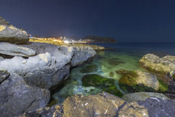 Scenic night view of the Mediterranean Sea at Costa Blanca, featuring rugged coastal rocks illuminated under a starry sky, reflecting tranquil lights and calm waters