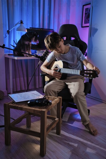 A young boy tuning his guitar in a cozy, blue lit room, with a microphone and sheet music nearby. A dog rests on a desk behind him, creating a serene atmosphere