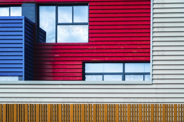 This image captures the vibrant and modern urban facade of a building, featuring a bold mix of red, blue, and white panels interlaced with clear windows, set against a clear sky
