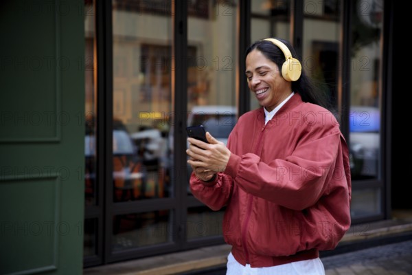 A man in a red jacket, wearing yellow headphones, smiles while looking at a smartphone. Captured outdoors against an urban backdrop, conveying joy and connectivity