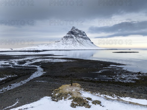 An unidentified man stands on a snow-dusted outcrop facing Kirkjufell, Iceland's iconic mountain, with icy shores and dramatic skies framing the serene and majestic winter landscape