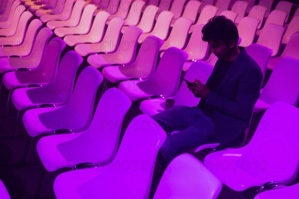 Indian man at a conference of AI in Amsterdam sits amid rows of vibrant purple lit chairs, deeply engaged with his smartphone. The scene captures solitude amidst innovation