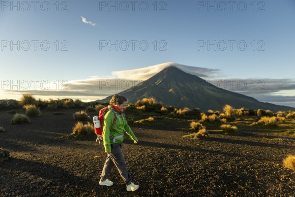 A woman hiker walks through the rugged terrain near Mount Taranaki, New Zealand, as the sun rises, casting a golden glow on the volcanic landscape. Perfect moment of outdoor exploration