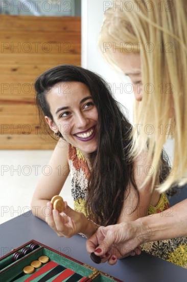 Mother and daughter smile and enjoy a friendly game of backgammon in a relaxed setting They interact over the board, holding playing pieces, showcasing social and leisure activities