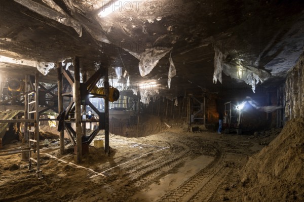 An expansive underground construction site featuring heavy machinery, scaffolding, and industrial lighting. Sandy terrain, tire tracks, and equipment show active work in progress