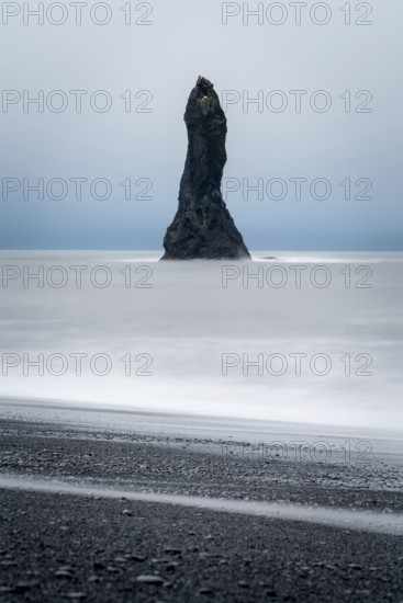 A dramatic sea stack rises from the ocean at Reynisfjara Beach in Iceland, surrounded by mist and black sand. A striking example of Iceland's unique winter landscape