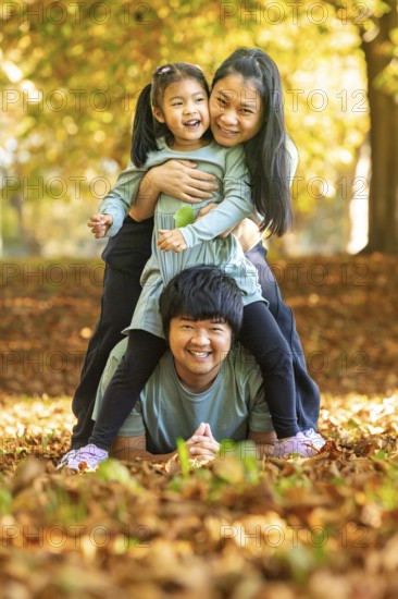 Asian family enjoying a playful moment outdoors in an autumn park. The child rides on the father's back, while the mother supports the child, surrounded by vibrant fall leaves