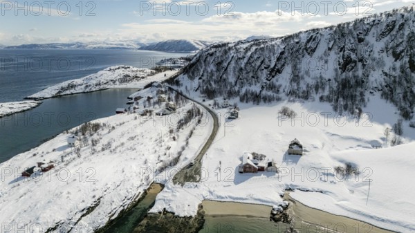 Aerial view of a serene Nordic landscape showcasing a snow-covered coastline, winding road, and scattered rustic houses against a backdrop of majestic snowy mountains, Botnhamn, Troms, Norway
