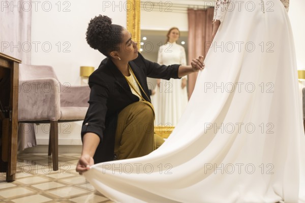 A seamstress carefully adjusts a flowing white bridal gown for a customer in a stylish bridal boutique. The focus is on meticulous attention to detail in dressmaking