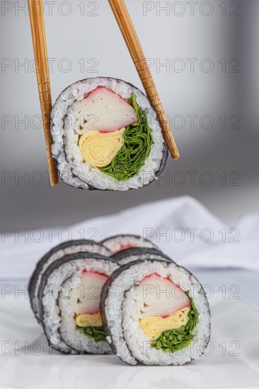 A close up view of sushi rolls with various colorful ingredients like crab, omelette, and greens wrapped in seaweed and rice in white background. Chopsticks hold one piece, showcasing its texture