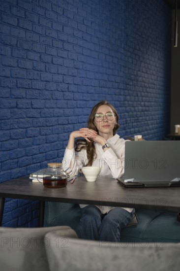 A woman with glasses sits at a cafe table with a laptop, tea cup, and teapot, against a stylish blue brick wall, taking a relaxing break from work