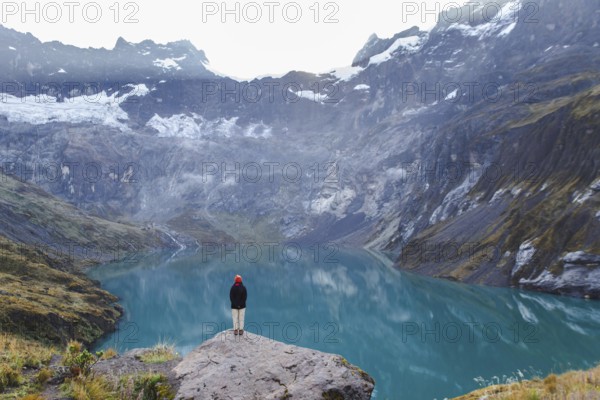 An Ecuadorian woman stands on a rocky ledge, overlooking the serene turquoise waters and majestic peaks of El Altar volcano in Ecuador, surrounded by misty clouds