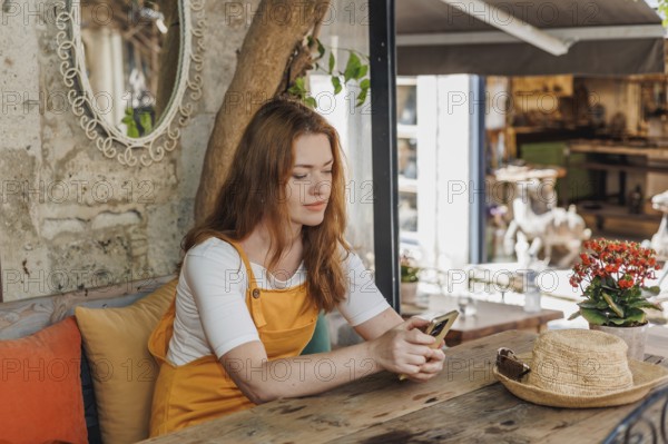 A mature woman tourist uses a smartphone while relaxing at a cafe in Cesme, Turkey. She enjoys her vacation, wearing orange and yellow attire