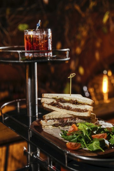 A gourmet setting features a stacked club sandwich with veal cheeks, cheese, and fresh greens, paired with a vibrant Negroni. Candlelight ambiance enhances the dining experience