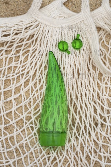 Top view of a green aloe vera gel tube and two green capsules placed on a white net bag, set against a sandy texture. Ideal for showcasing natural skin and hair care products