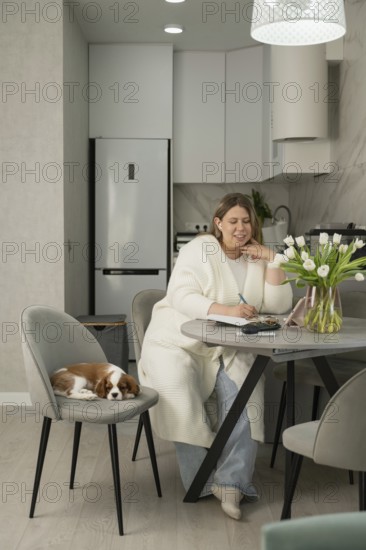 A woman sits at a modern kitchen table, writing in a notebook. Nearby, a small dog rests on a chair. Tulip vase adds elegance to the cozy, light-filled space