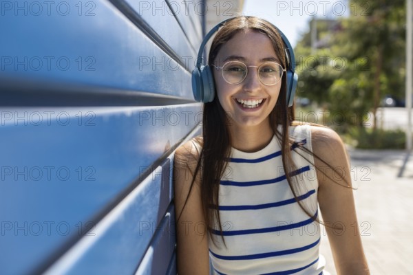 Smiling young woman with headphones enjoys music while leaning against a blue wall in an urban setting, embodying an active and vibrant city lifestyle