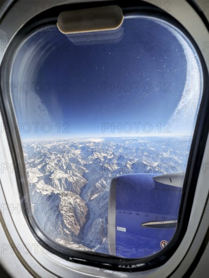 Stunning aerial view of a vast snow-covered mountain range as seen through an airplane window. The image captures the serene beauty of the landscape from high above the clouds