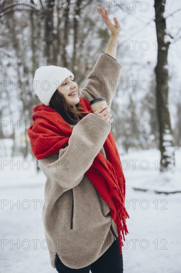 A cheerful young woman smiles as she lifts her hand in a snowy park, dressed warmly with a red scarf, white beanie, and holding a coffee cup