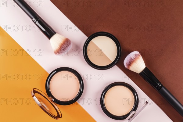 Flat lay of makeup brushes and face powders on a vibrant tricolor background. The contrasting orange, white, and brown surfaces highlight the beauty products