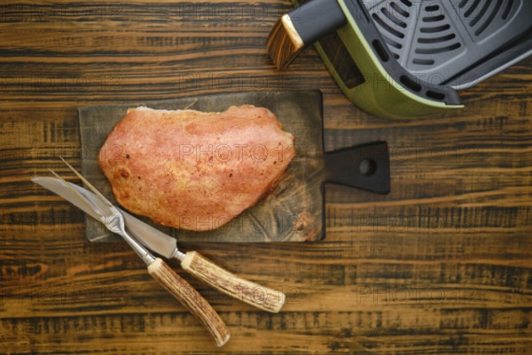 A piece of roasted pork meat on a wooden cutting board. Kitchen knife and fork are placed next to the fillet. An air fryer is seen on the side of the board. Photo with shallow depth of field