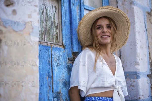 A smiling woman in a straw hat leans against a rustic blue door in Trypiti, Milos Island, Greece. The setting exudes a relaxed summer vibe with a touch of Mediterranean charm