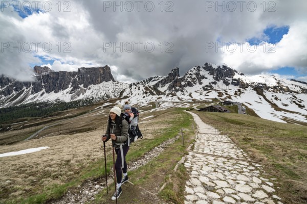 A mother and her young son enjoy a hike at Passo Giau in the Dolomites, Italy. They walk along a scenic stone path surrounded by snow-capped mountains, creating a striking alpine adventure atmosphere