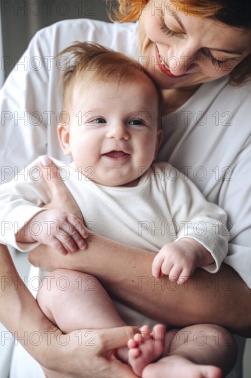 Loving mother cradles smiling newborn baby girl at home, both wearing white clothes. The baby's joyful expression enhances the warmth of their shared moment