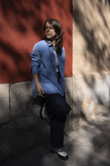 A stylish woman in a blue shirt leans against a red and beige wall, gazing into the distance. The interplay of light and shadows creates an artistic and contemplative atmosphere in the urban setting