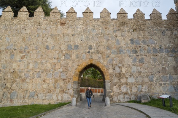 An Asian woman walks through an arched entrance of a medieval city wall in Avila, Spain, surrounded by historic stone architecture, showcasing the blend of history and culture