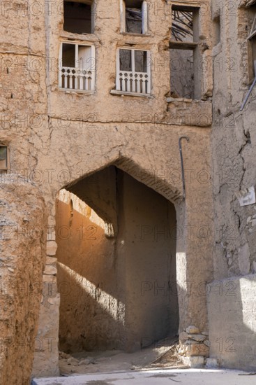 A textured, sunlit archway casts golden hues across the crumbling facades of traditional mud-brick buildings in Al Hamra, Oman, showcasing the historic architecture of this abandoned village